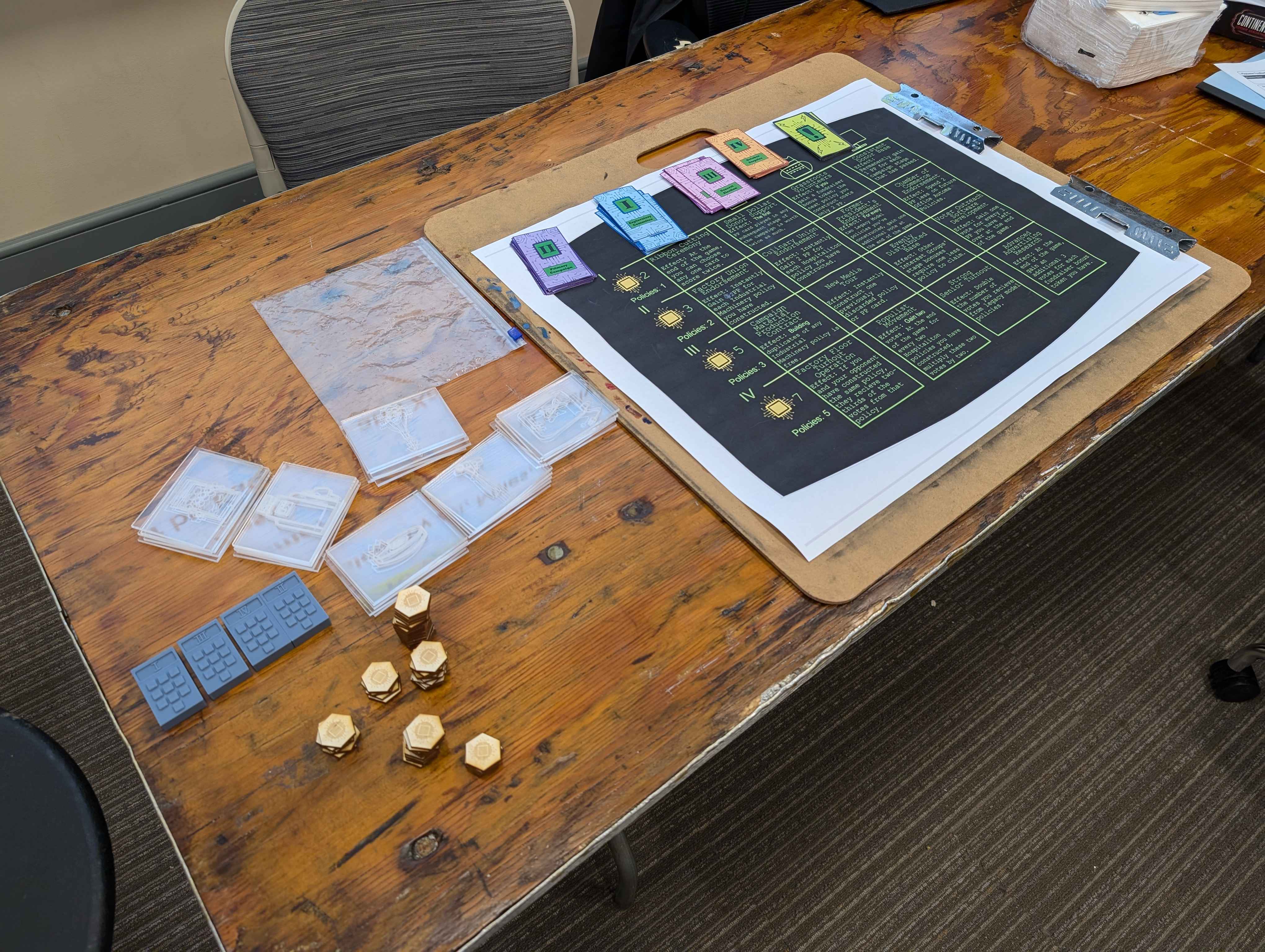 A photograph of the fabricated materials for the board game sitting on a wooden table. The table has clear acrylic tiles with laser-etched images, laser-cut wooden tokens, printed cards and a printed game board, and 3D printed calculator tokens.